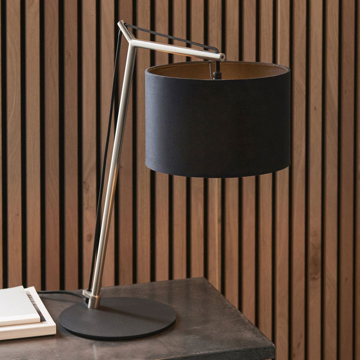 Black and gold table lamp on a dark gray table with a wooden slatted wall behind it.