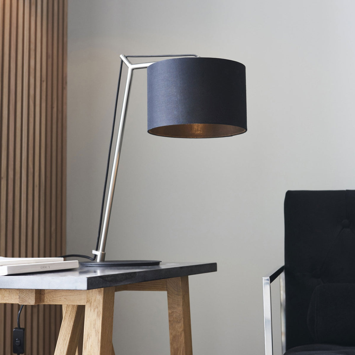 Black and gold table lamp on a dark gray table next to a black chair, with a wooden wall.