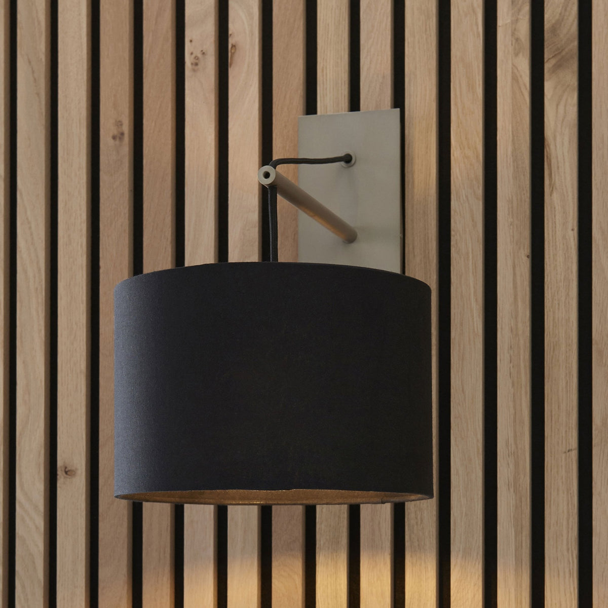 Black drum shade wall light with a wooden arm and a light grey backplate on a wooden slat wall.