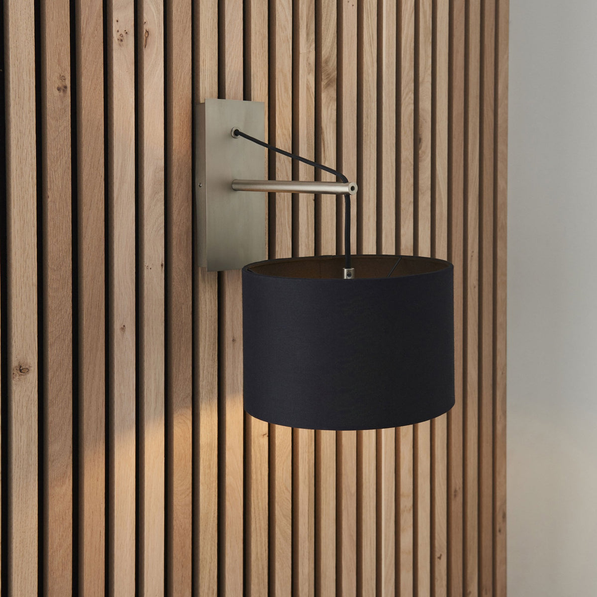 Black drum shade wall light with a brushed steel arm and backplate on a wooden slat wall, casting light downwards.