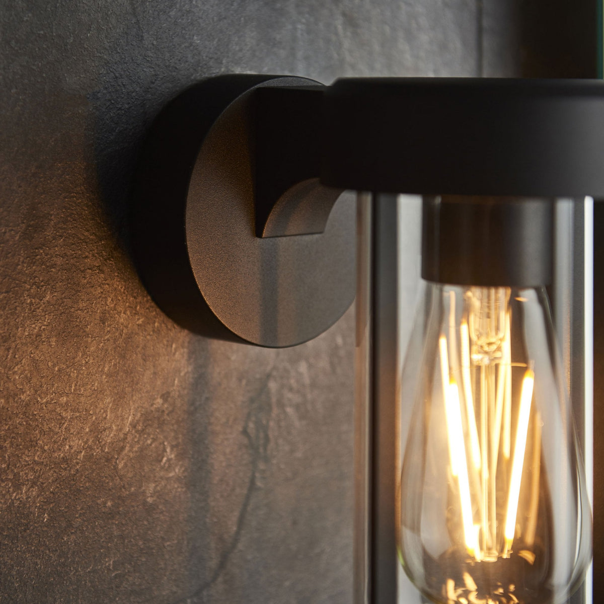 A close-up of a black modern outdoor wall light with an exposed Edison bulb, on a dark tiled wall.