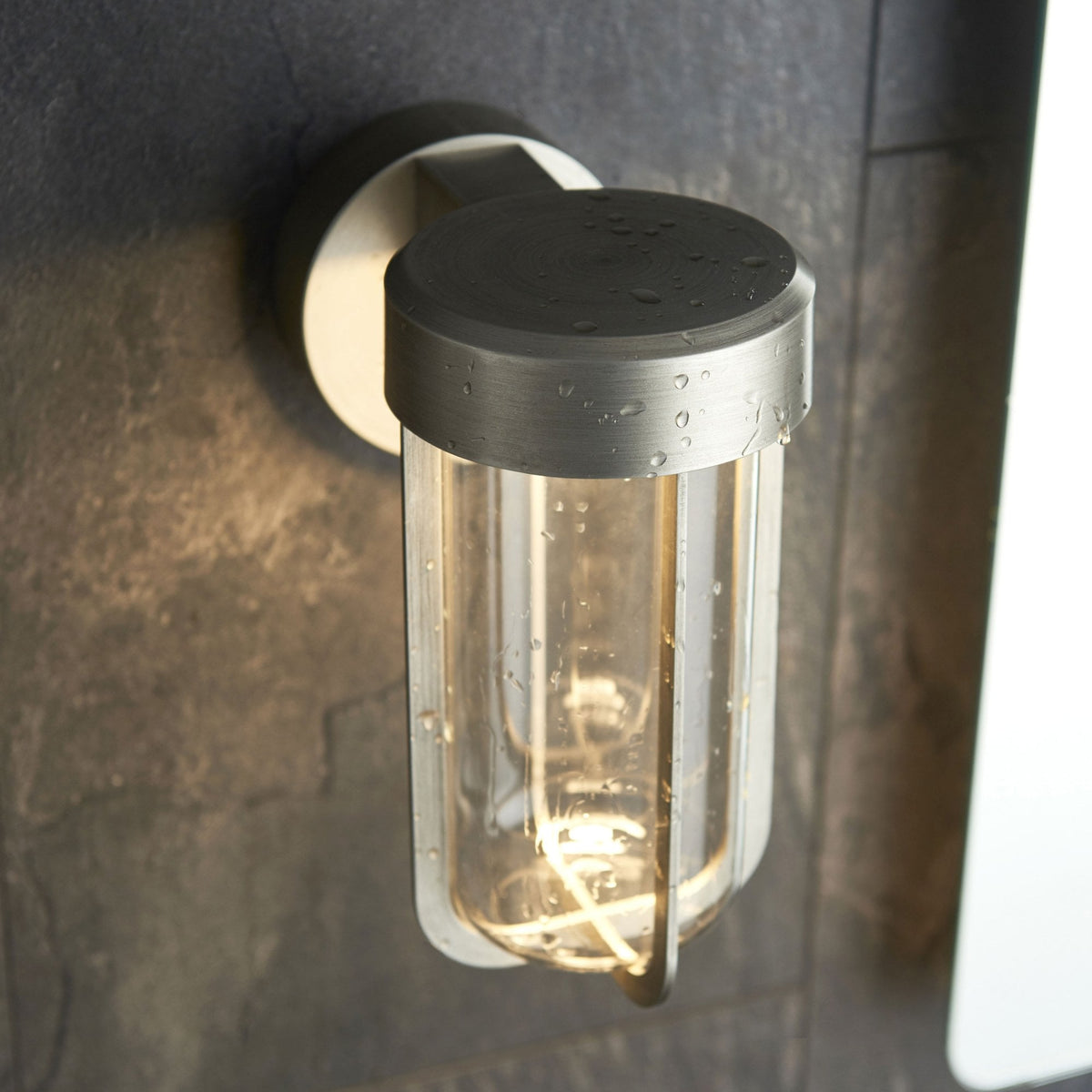 Brushed steel outdoor wall light with clear glass and water droplets, mounted on a dark, textured wall.