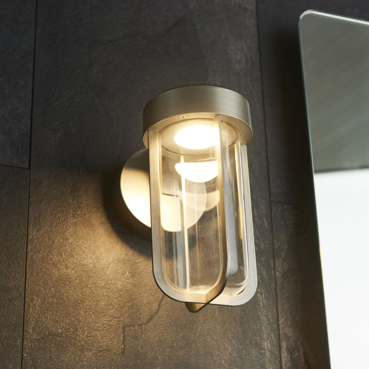 Contemporary outdoor wall light with a brushed metal finish and clear glass, providing subtle illumination on a dark wall.