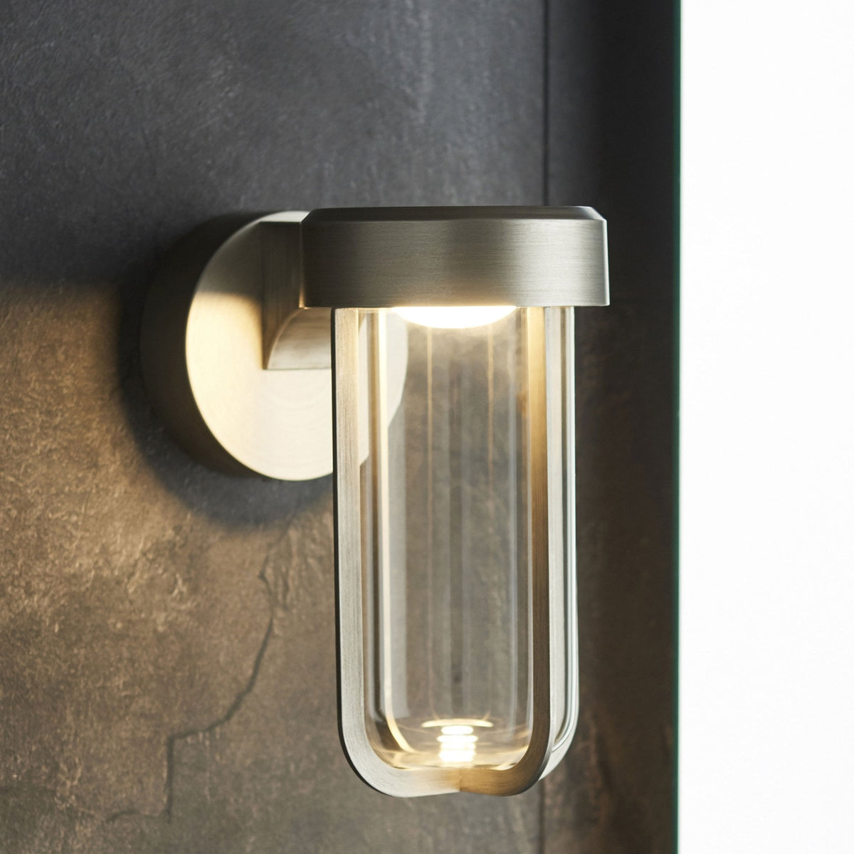 Sleek outdoor wall light with a brushed steel finish and clear glass, casting light on a dark, textured surface.