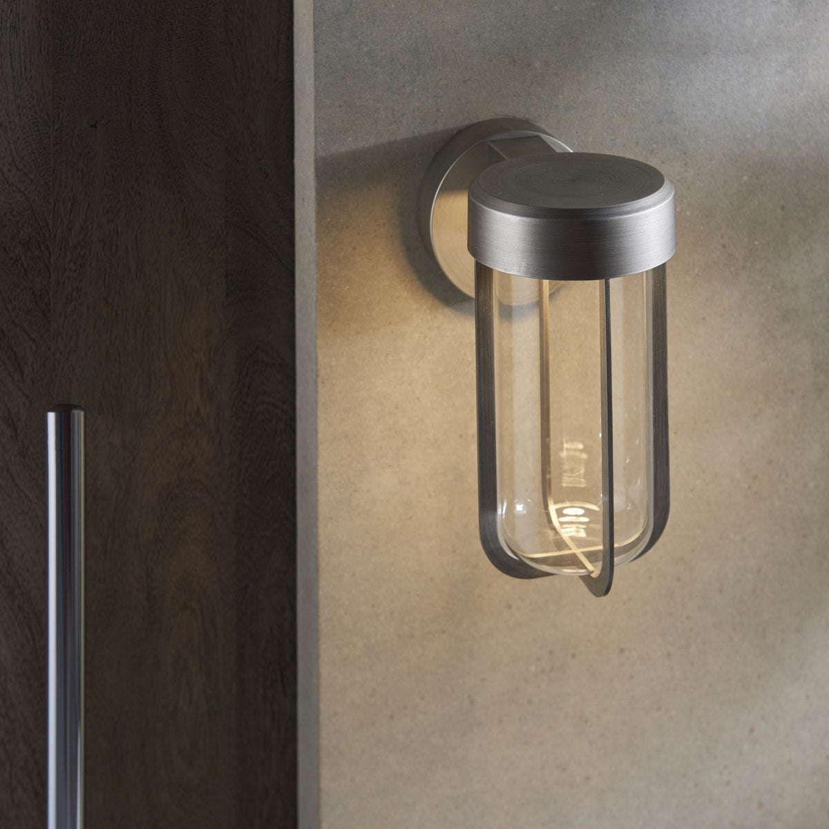 Modern brushed steel outdoor wall light with clear glass, illuminating a subtle, textured wall in an outdoor setting.
