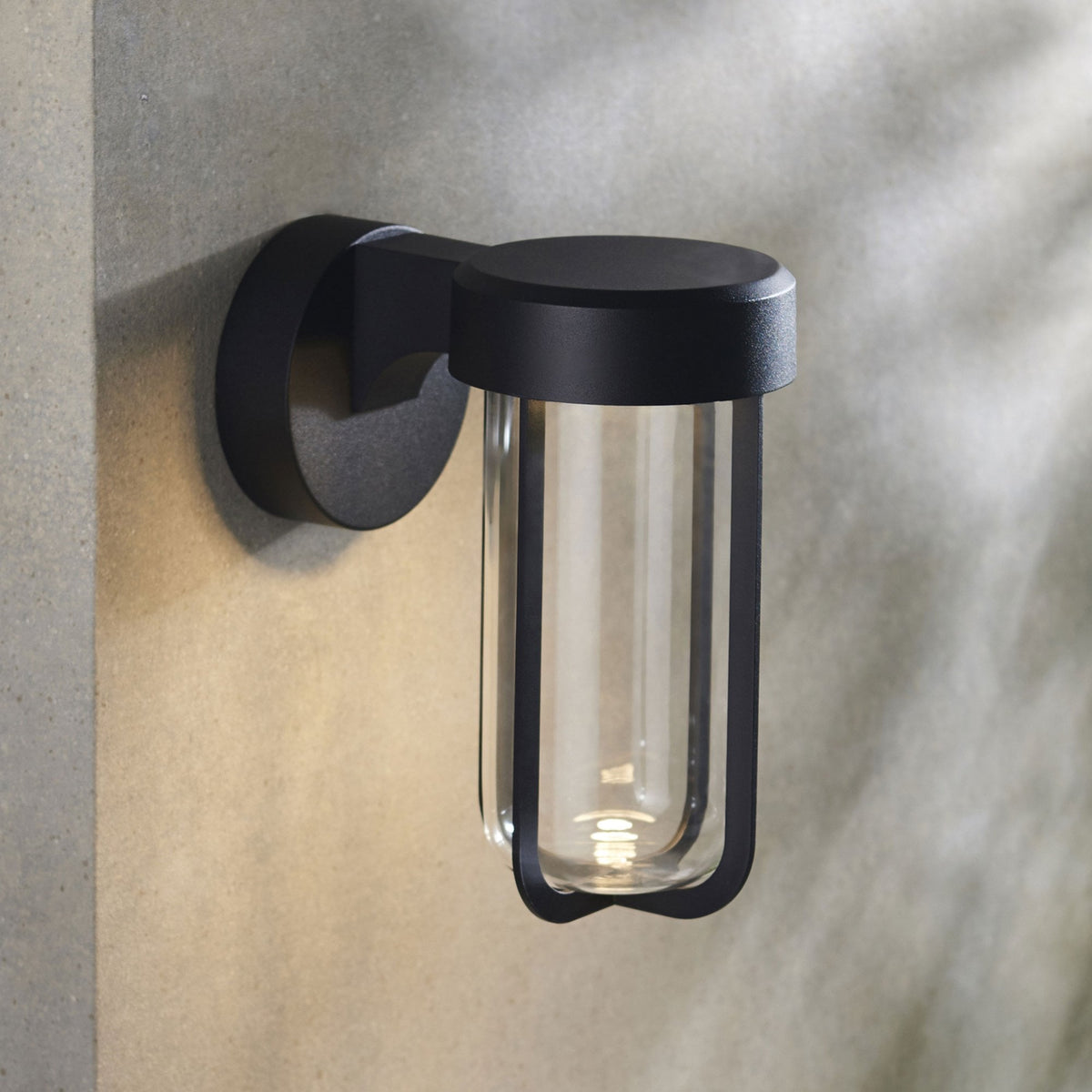 A close-up shot of the matte black, modern wall sconce with a clear glass shade, emitting warm light.