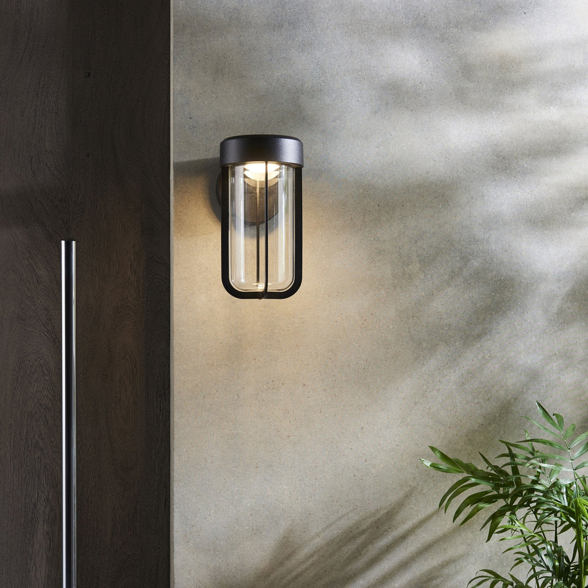 A modern outdoor wall sconce with a clear glass cylinder and black frame, illuminating a textured wall.