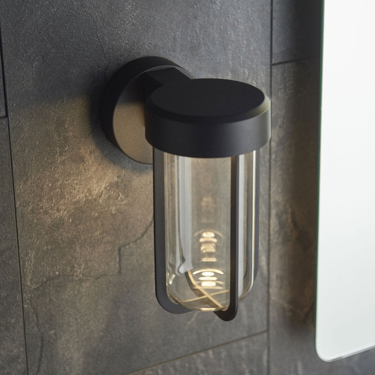 A black modern wall sconce with a clear glass cylinder and an exposed filament bulb on a dark wall.
