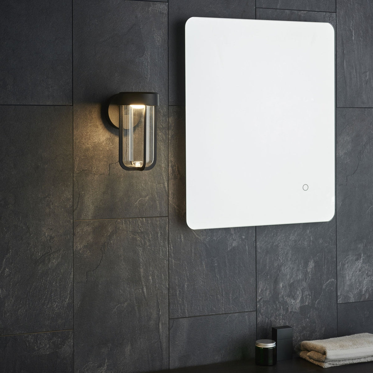 A black outdoor wall light next to a rectangular mirror, set against a dark gray tiled wall.