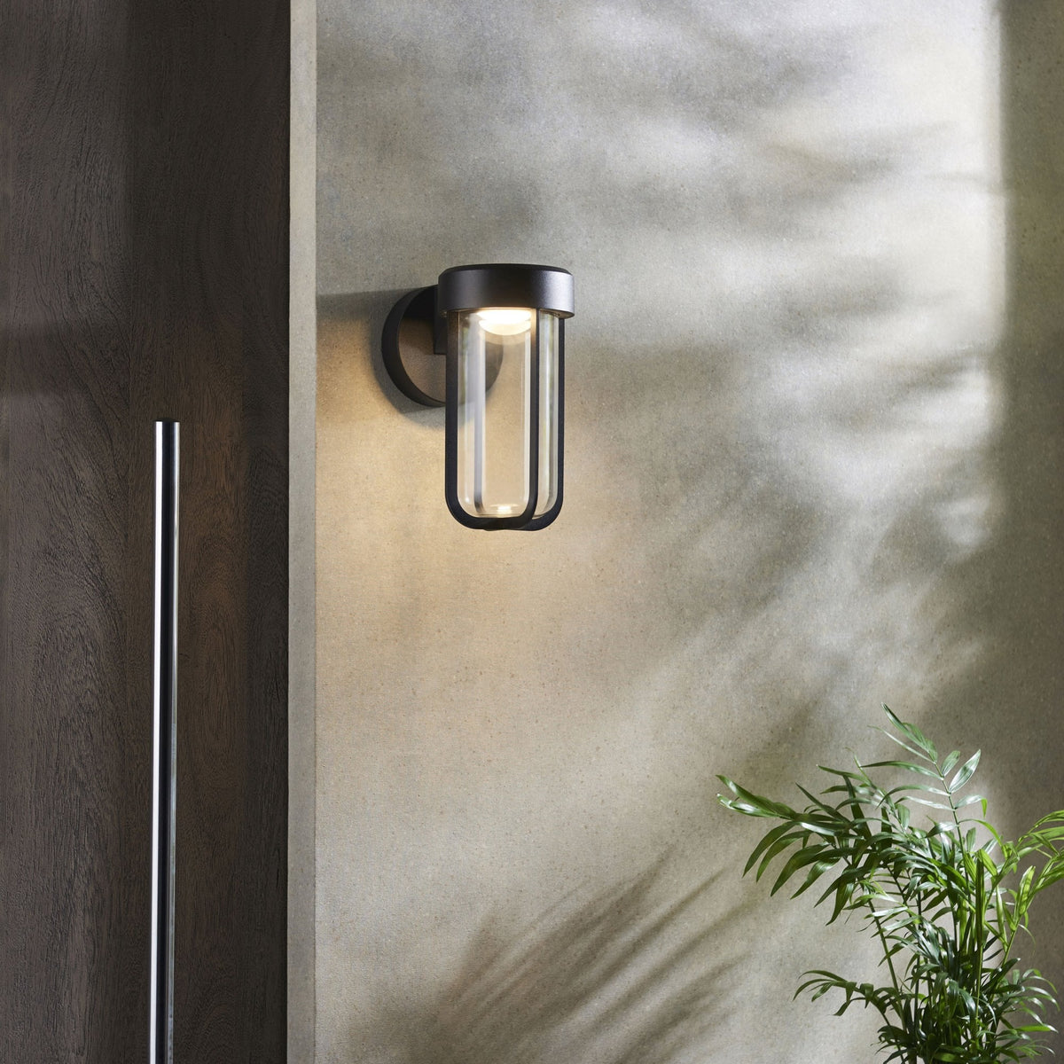A modern black wall light with a clear glass cylinder casts warm light on a textured outdoor wall.
