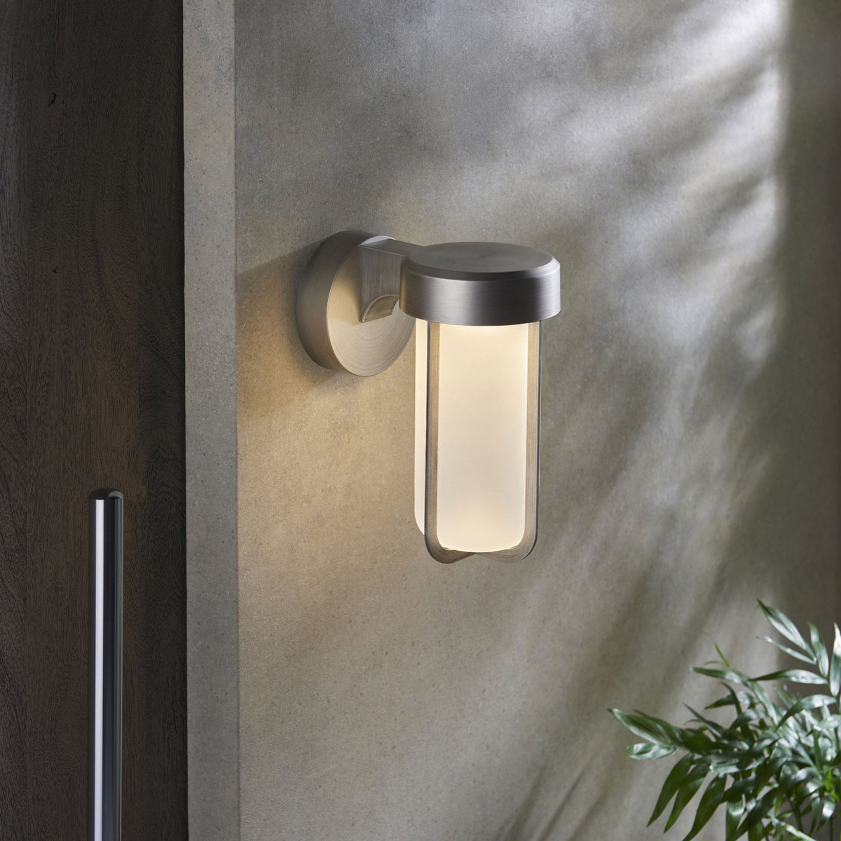 A contemporary outdoor wall light with a brushed metal finish and a glowing frosted glass shade on a light grey wall.