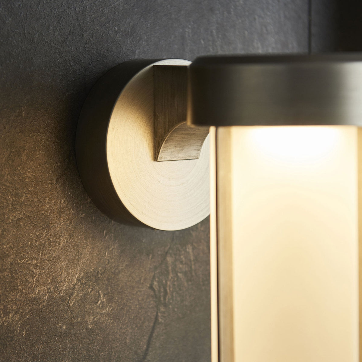 Close-up of a contemporary outdoor wall light's brushed metal base and glowing frosted glass on a dark stone wall.