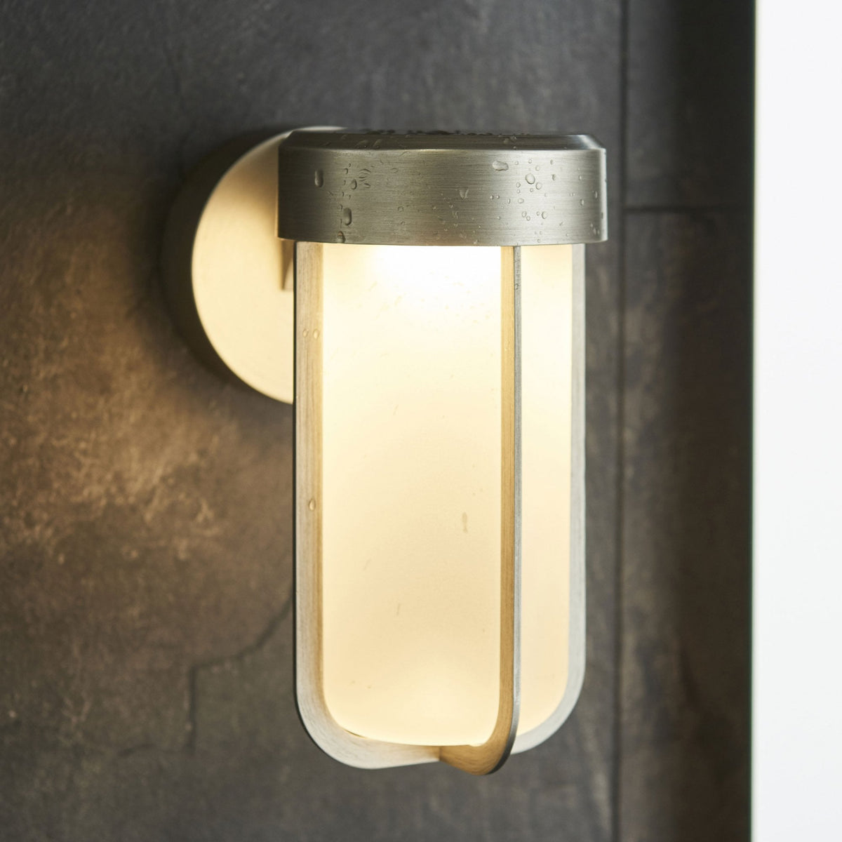 A modern outdoor wall light with a brushed steel finish and frosted glass diffuser with water drops.