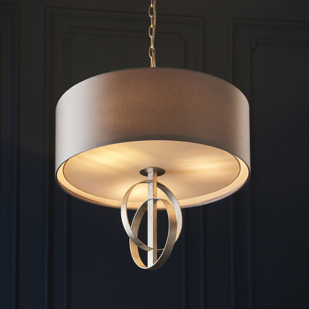 A beige drum shade pendant light with intertwined metallic rings, illuminated against a dark blue wall.