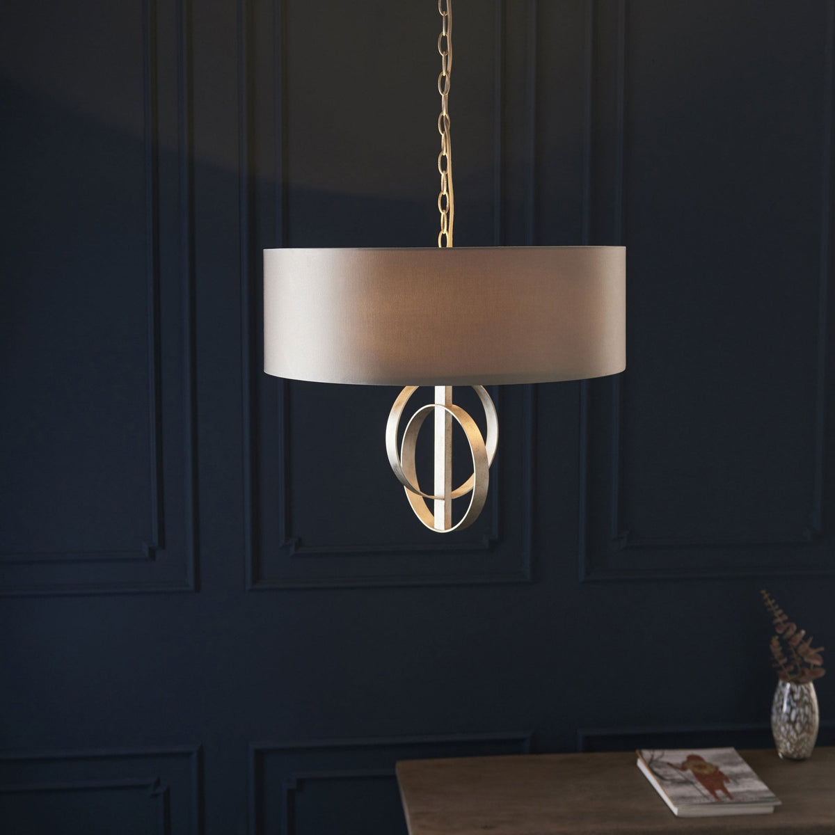 A beige drum shade pendant light with metallic rings above a wooden table in a dark room.