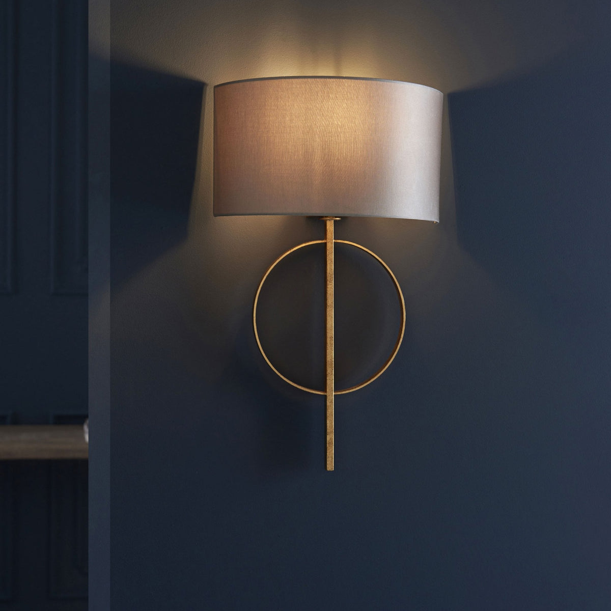 A modern wall sconce with a gold finish and a grey drum shade illuminates a dark blue wall.