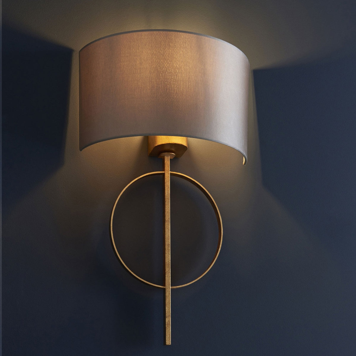 A close-up of a wall light, featuring a gold circular base and a grey fabric drum shade on a dark wall.