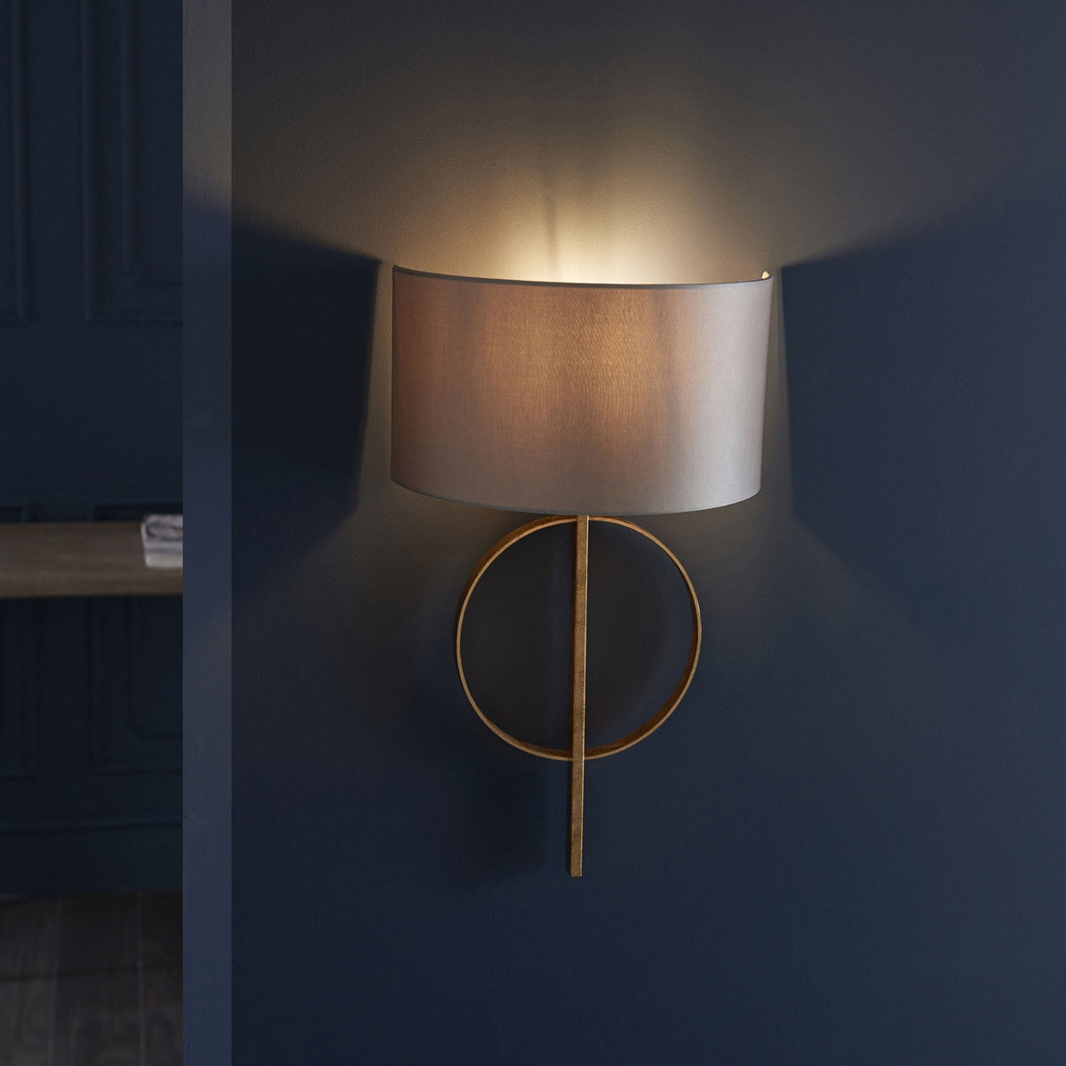 A contemporary gold and grey wall sconce emits a warm glow against a dark blue paneled wall.