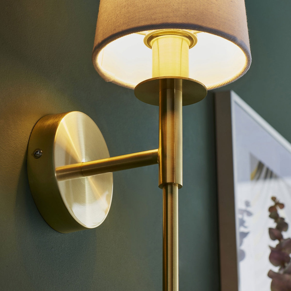 Close-up of a brass wall light, revealing the texture of its fabric shade and warm light against a dark green wall.