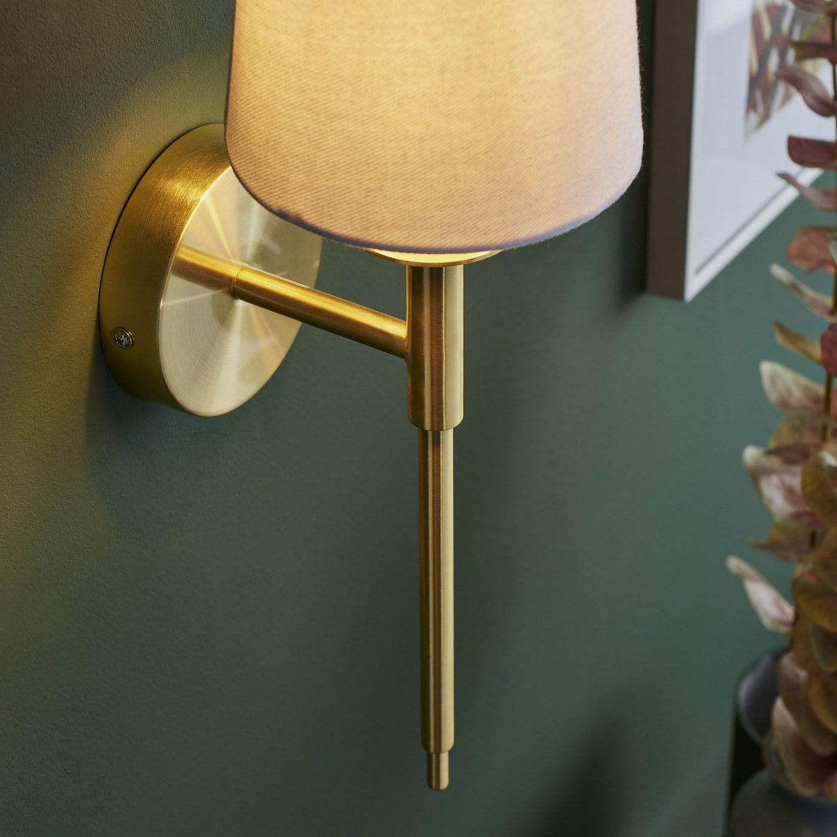 A brass wall light with a fabric shade, casting a soft glow on a deep green wall.