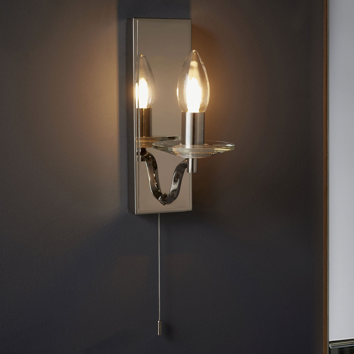 Polished chrome wall light with a clear glass candle holder and a pull cord switch, mounted on a dark grey wall.