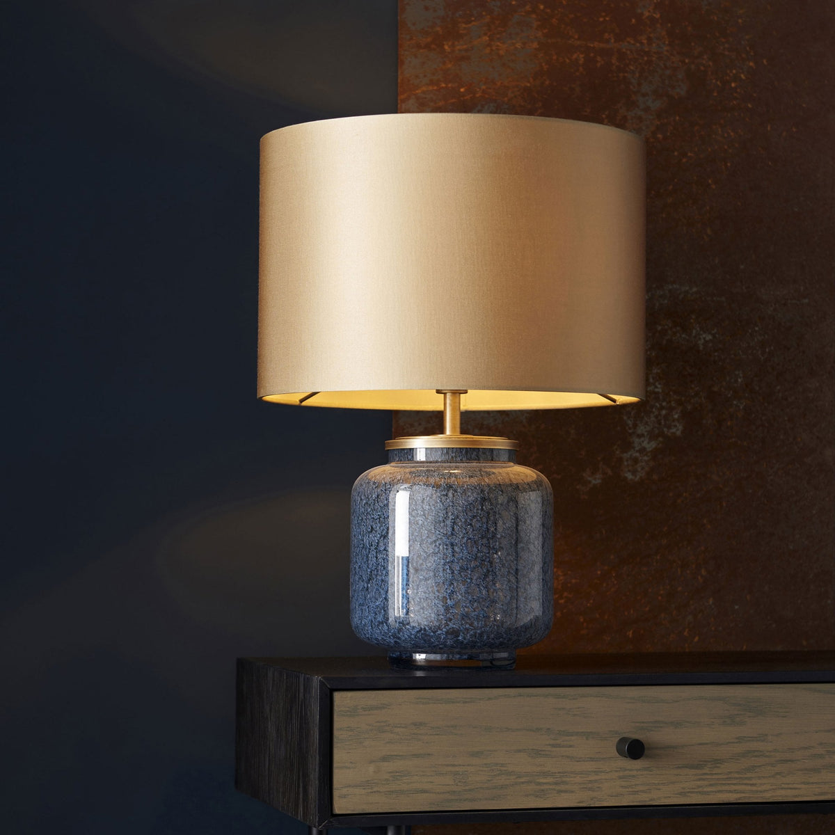 A blue glazed table lamp with a brass base and a gold lampshade, on a dark wooden console table.