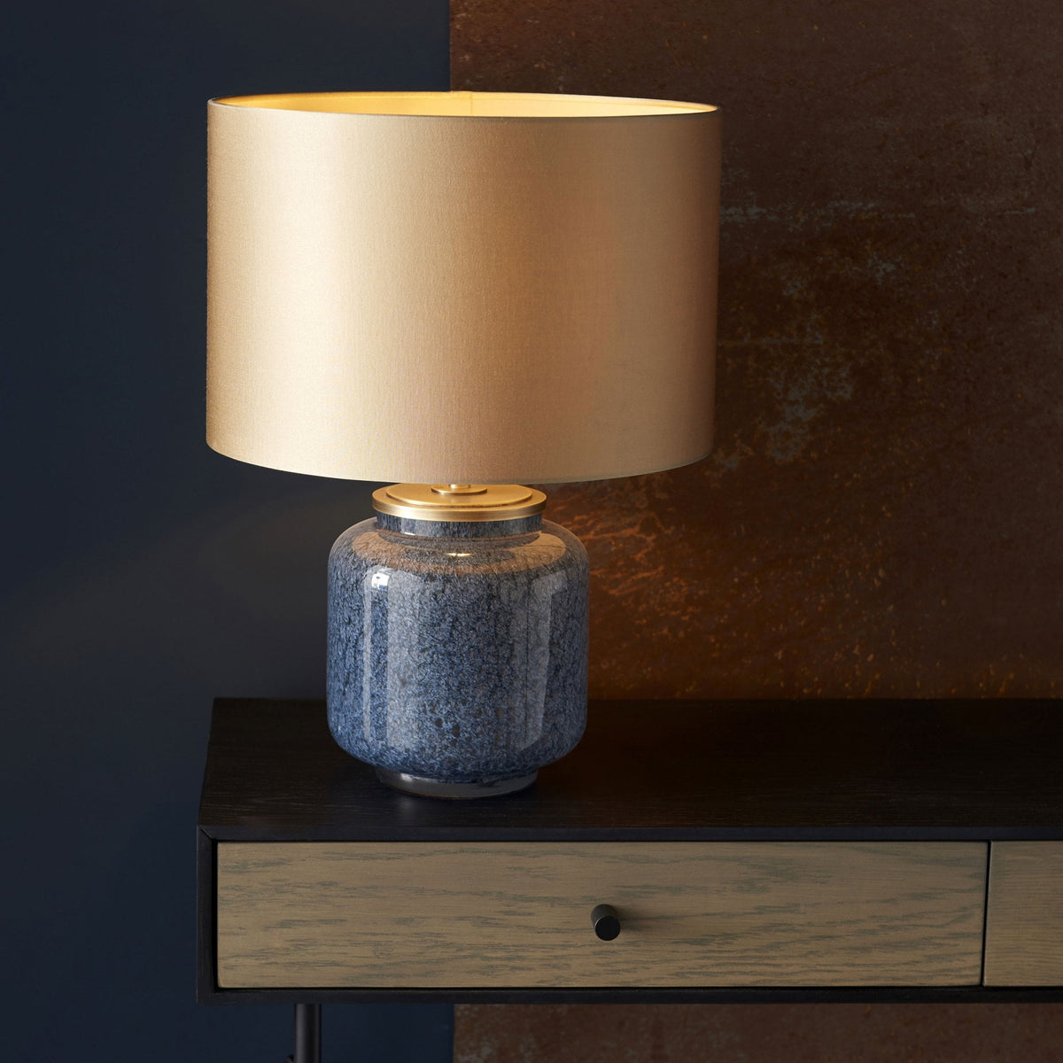 A blue glazed table lamp with a brass base and a gold lampshade, on a dark wooden console table.