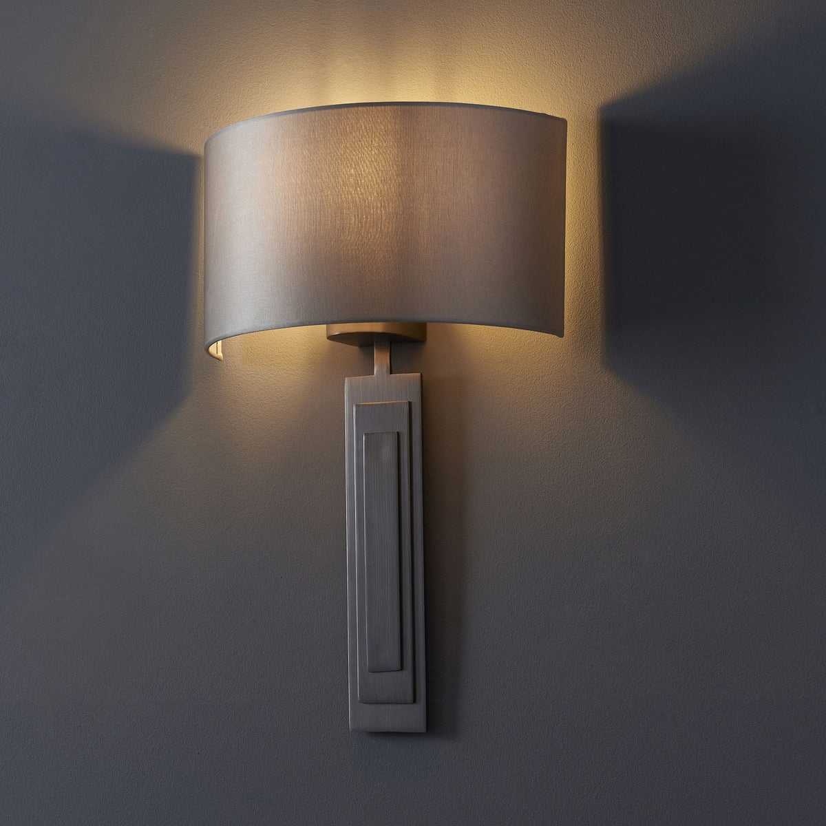 A grey wall light with a rounded shade casts a warm glow upwards and downwards on a dark grey wall.