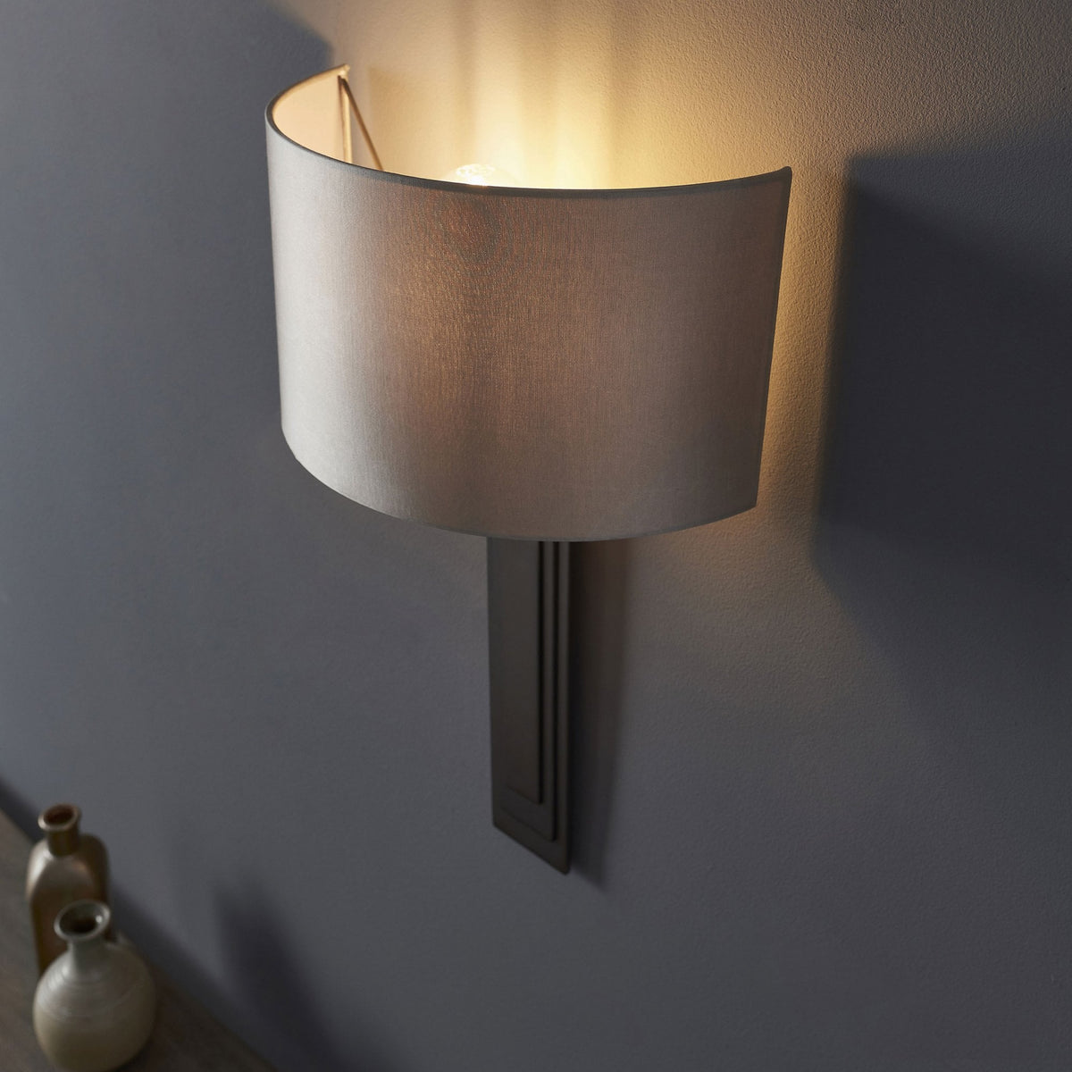 A grey wall light with a curved shade illuminates a dark grey wall, next to two small decorative vases.