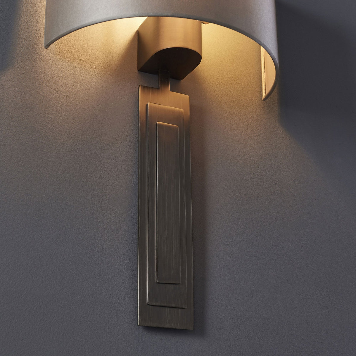 A wall-mounted light fixture with a curved grey shade and a detailed rectangular bronze base, emitting light.