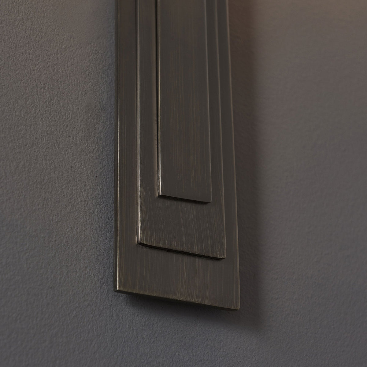 A close-up of the textured, rectangular base of a bronze wall light, featuring a subtle layered design.