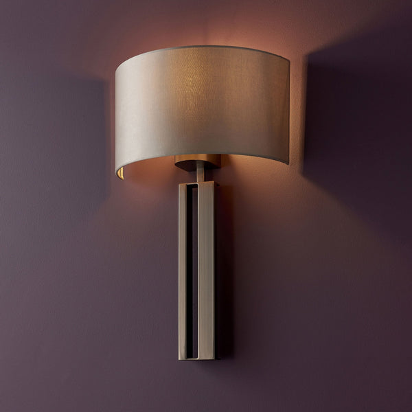 A modern wall light with a rectangular brass base and a cream fabric shade, emitting warm light.