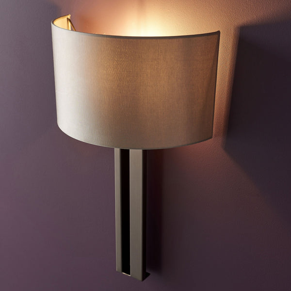 A modern wall light with a rectangular brass base and a cream fabric shade, emitting warm light.