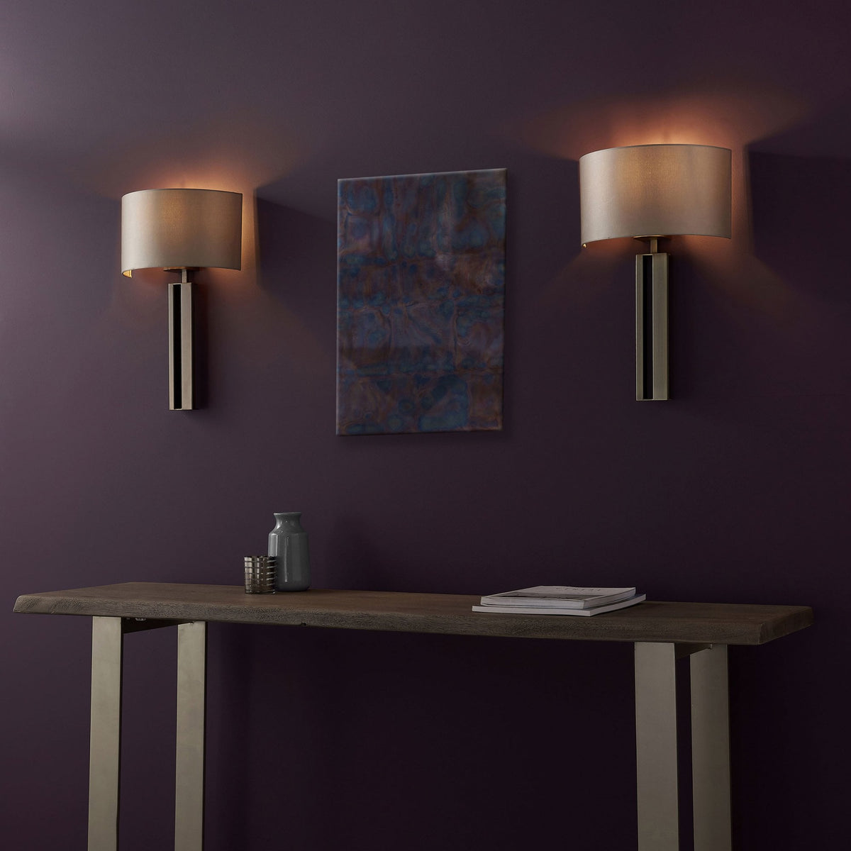 Two modern wall lights flank abstract art above a console table with decorative items on a dark wall.