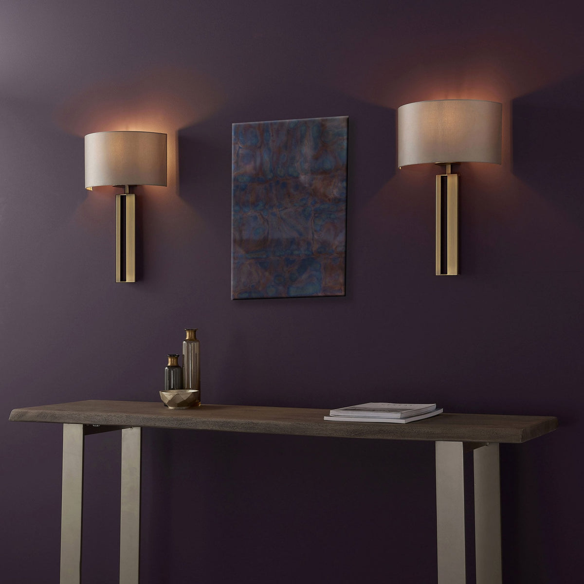 Two brass wall lights flanking an abstract art piece above a console table against a purple wall.