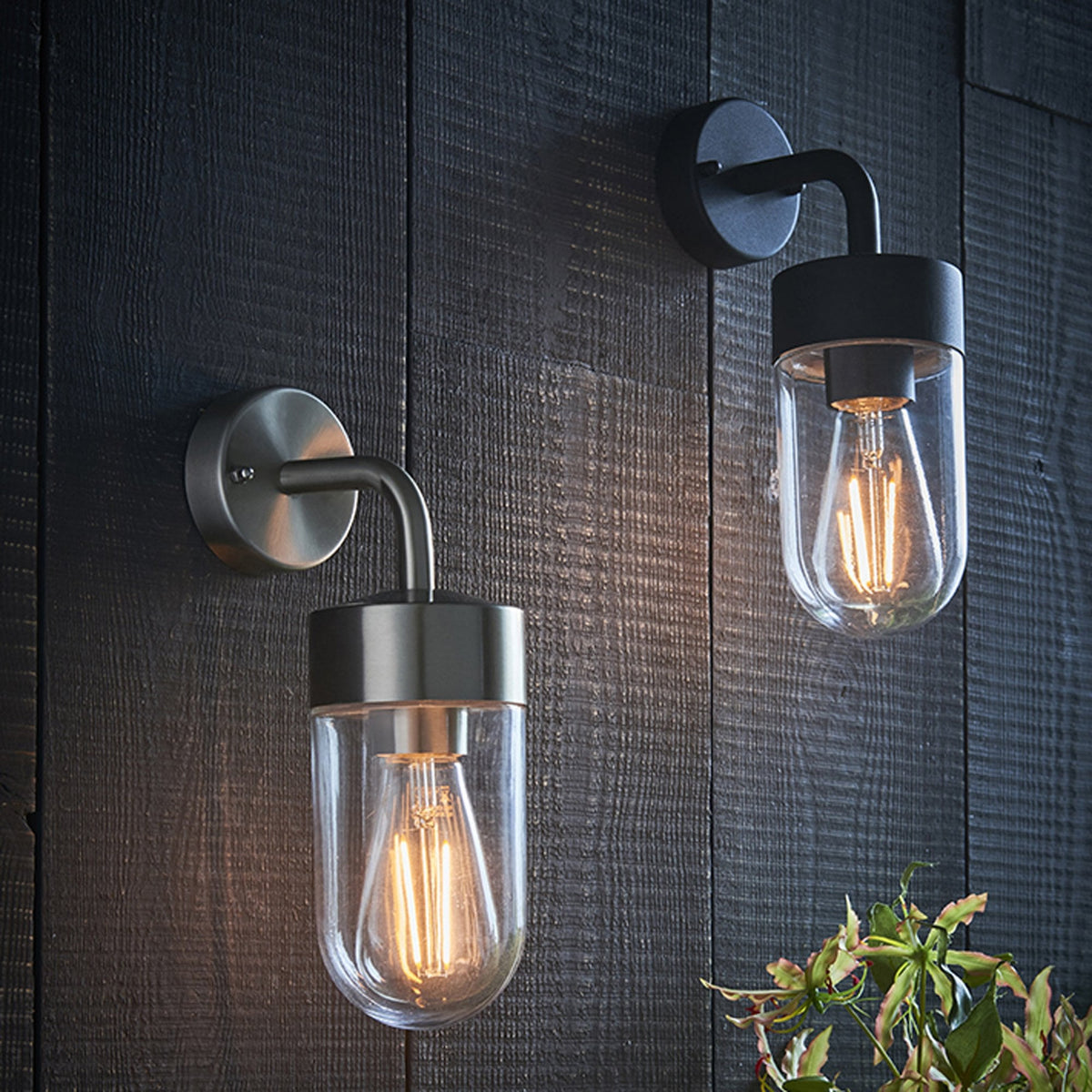 Two outdoor wall lights, one black and one antique brass, with clear glass shades on a dark wooden wall.