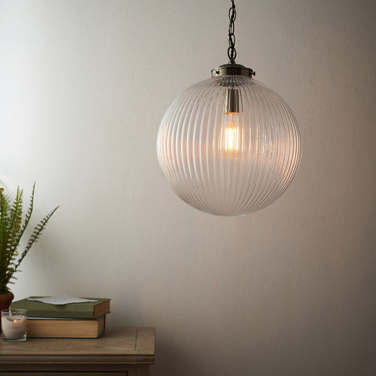 Ribbed glass globe pendant light with a warm glow, hanging above a wooden side table with books and a plant.