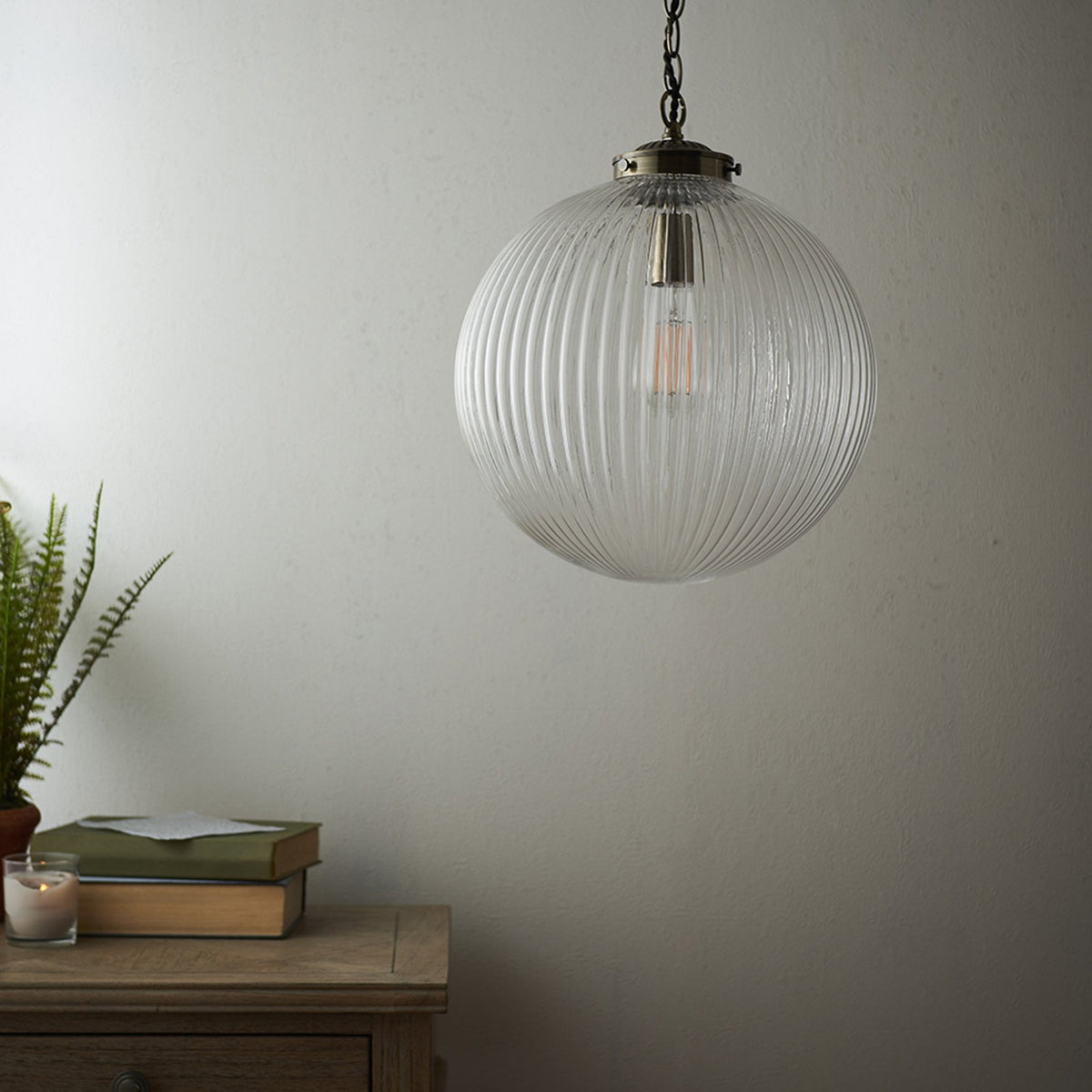 Ribbed glass globe pendant light with a warm glow, hanging in a room with a wooden side table and plant.