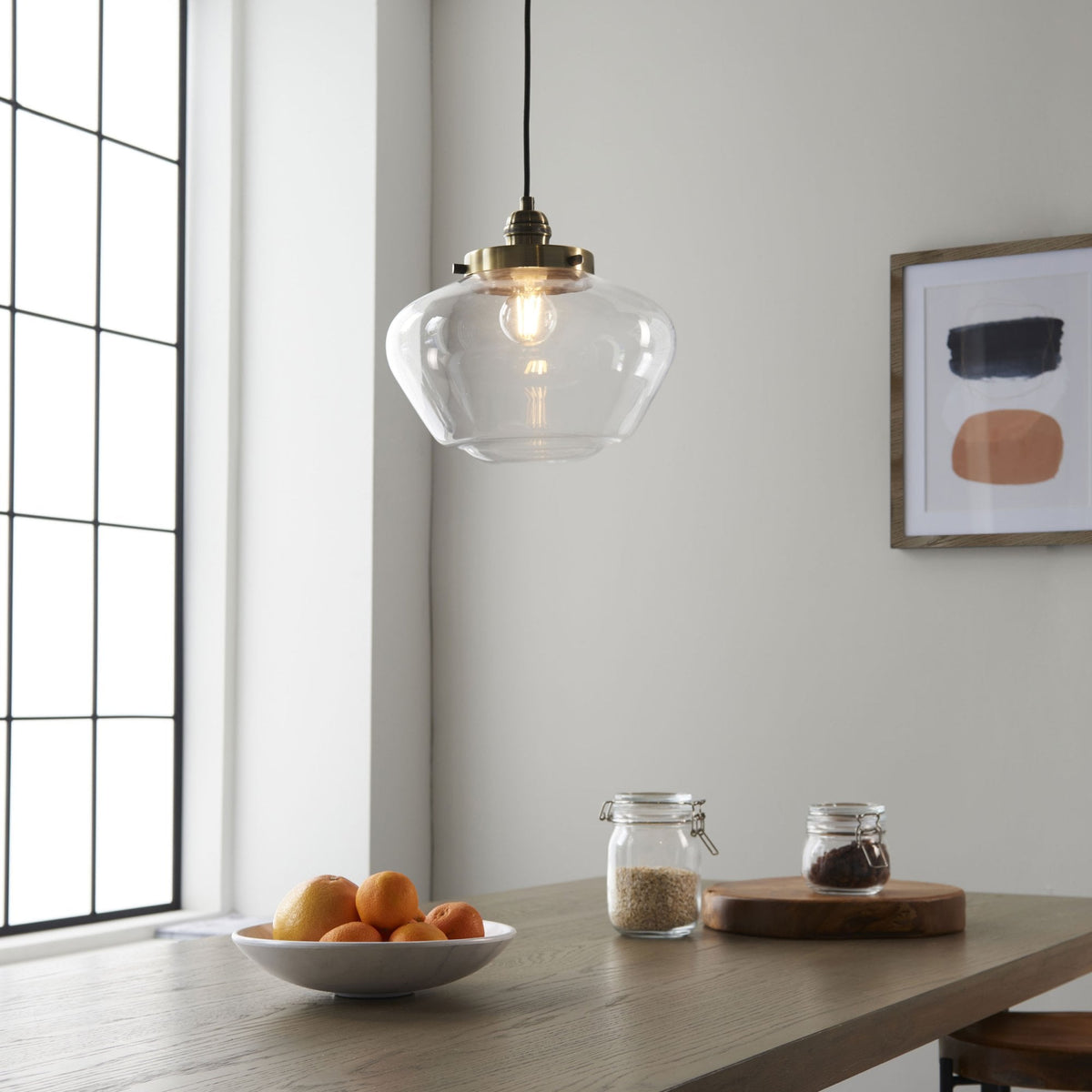 Clear glass pendant light illuminating a dining area with a wooden table, fruit bowl, and window.