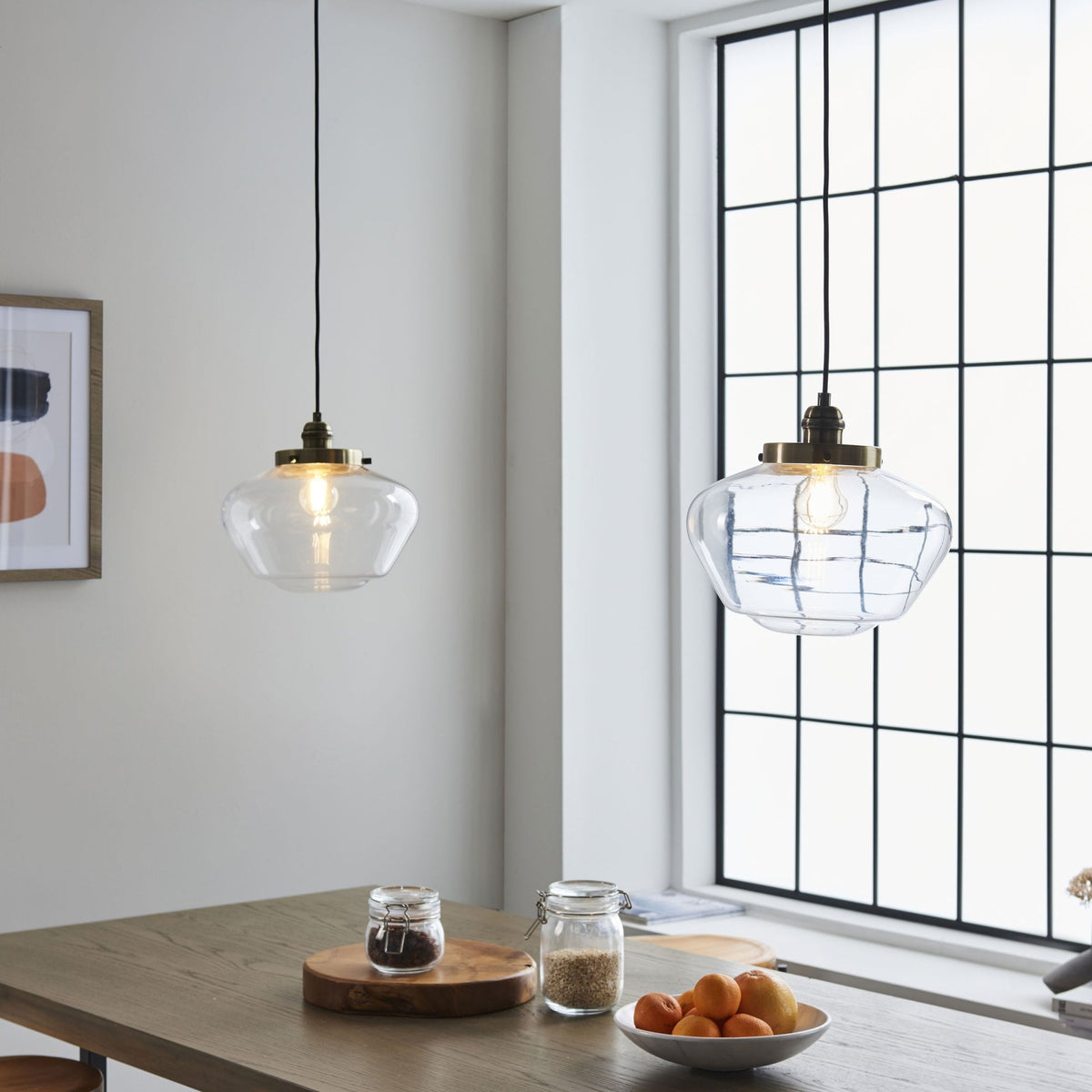 Two clear glass pendant lights hanging above a wooden table next to a large window.