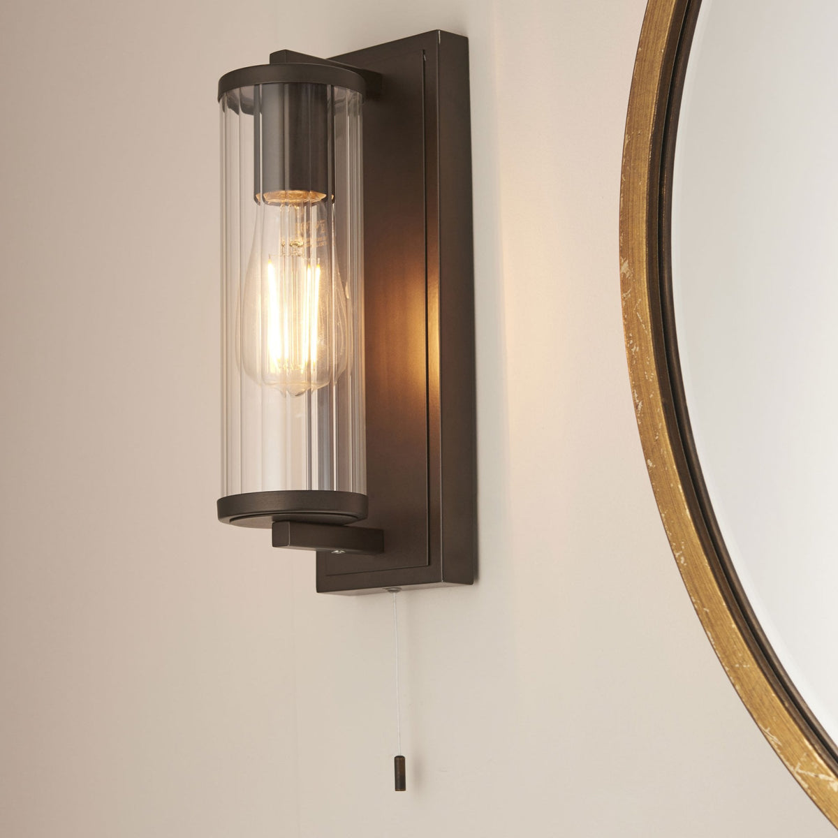 Bronze wall light with a clear ribbed glass shade and exposed filament bulb, next to a round mirror.