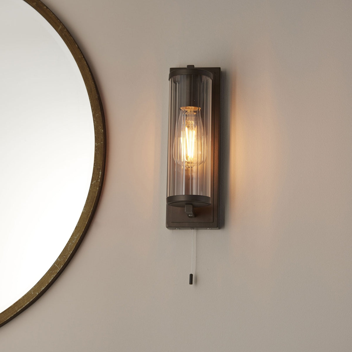 Bronze wall light with a clear ribbed glass shade and exposed filament bulb, next to a round mirror.