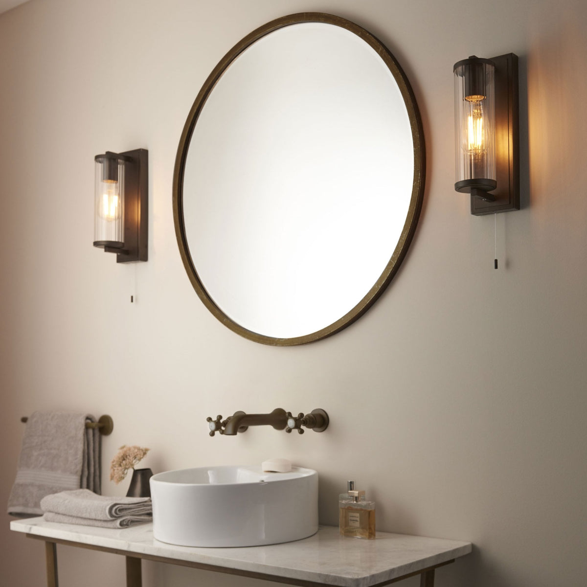 Two bronze wall lights with clear ribbed glass shades flanking a round mirror above a bathroom vanity.