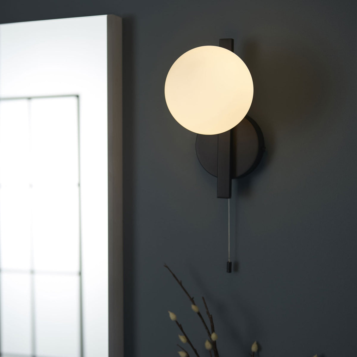 Black wall light with a round, white globe shade and a pull cord switch, mounted on a dark gray wall.