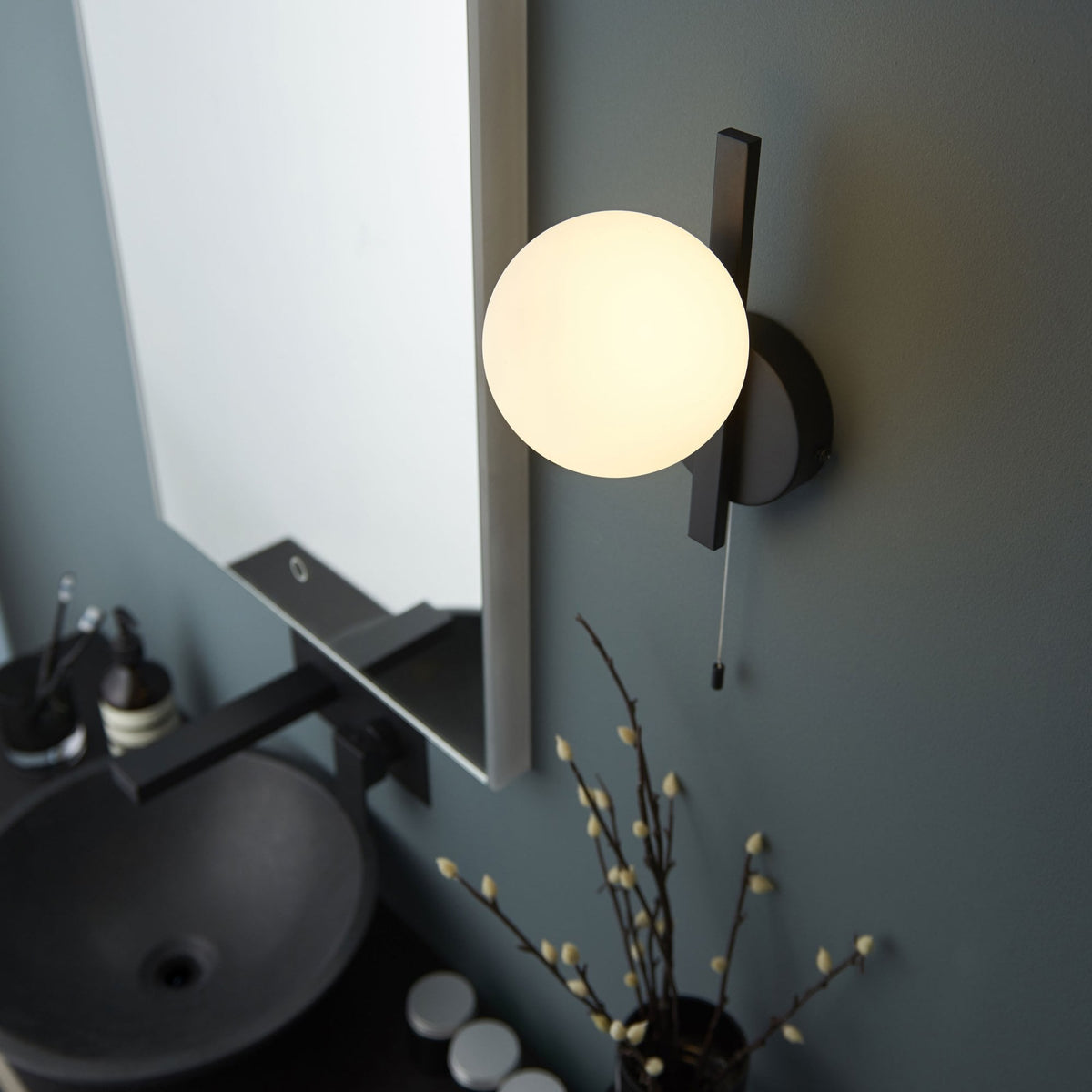 Black wall light with a white globe shade next to a mirror, black sink, and decorative branches.