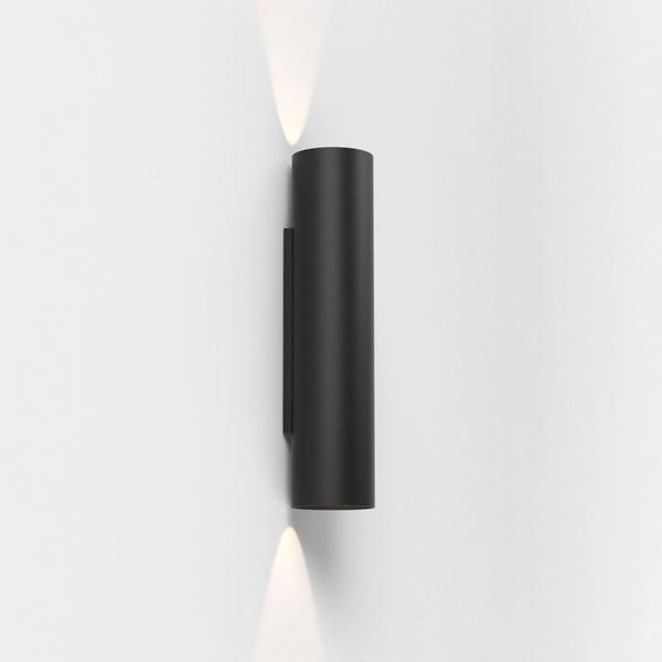 Black cylindrical wall light with light beams projecting upwards and downwards on a white wall