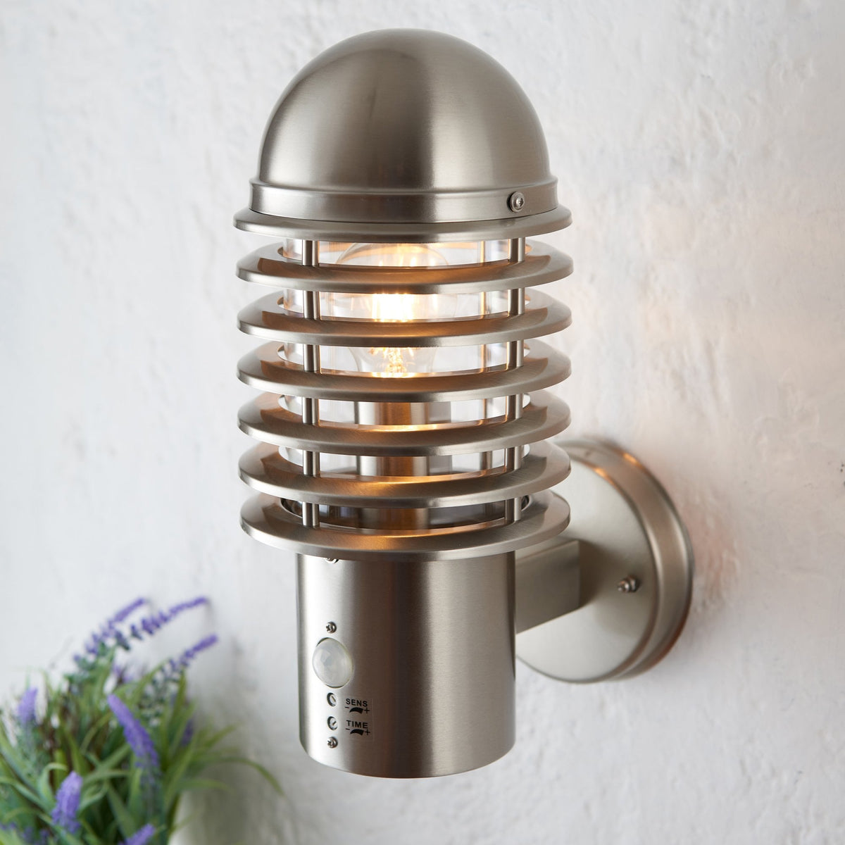 Brushed stainless steel outdoor wall light with a motion sensor and a warm glowing bulb.