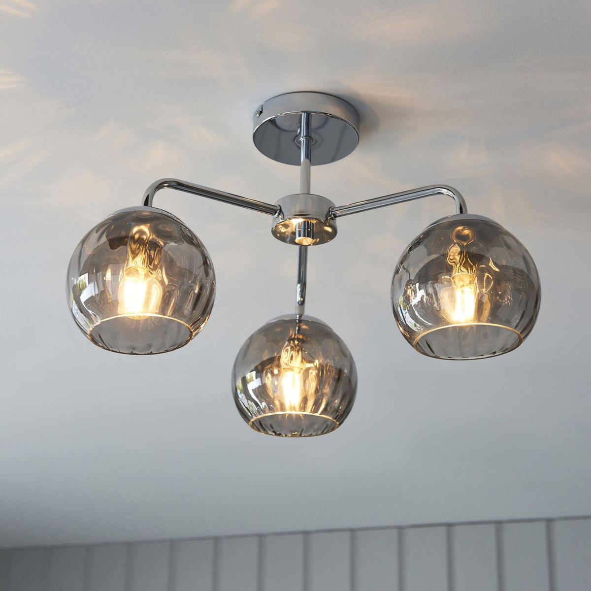Chrome ceiling light with three smoked glass shades, casting warm light patterns on the white ceiling.