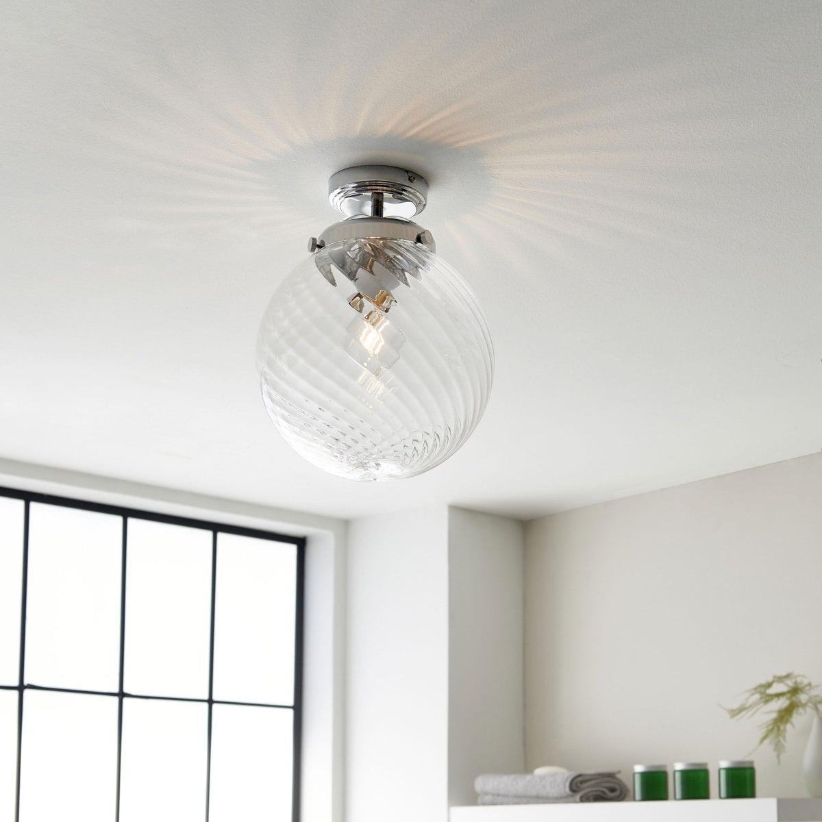 Clear glass globe ceiling light with a chrome base and a swirling pattern, casting light in a room with a window.