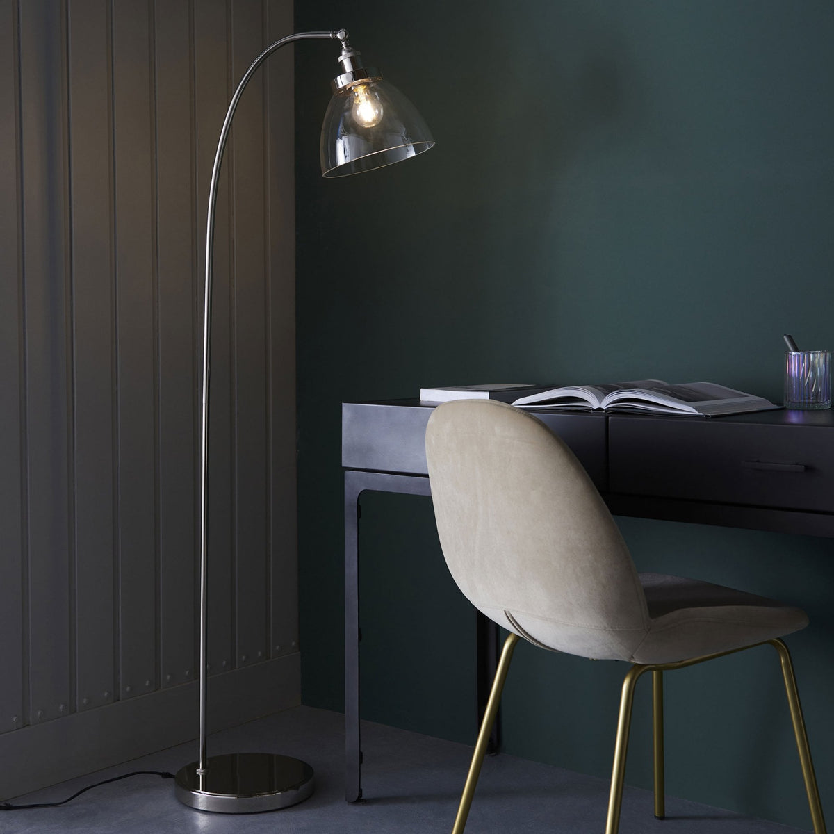 Floor lamp with a curved silver stand and clear glass shade next to a dark desk and a beige chair.